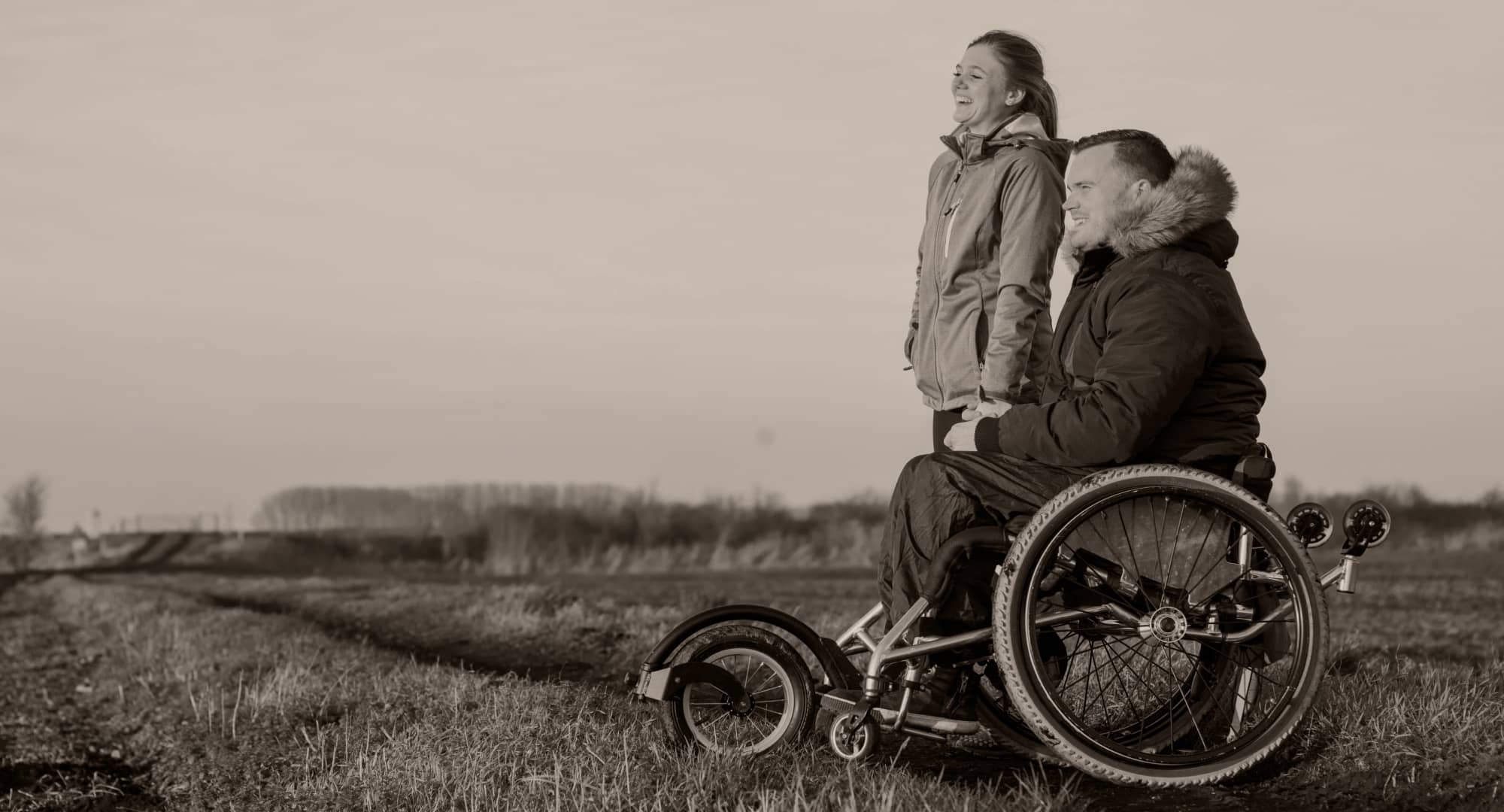 Woman stands holding hands with man in wheelchair as they share a smile looking across open fields